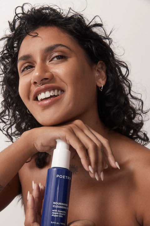 POETRY Skincare. Woman with brown curly hair, brown eyes, brown skin, nose ring smiling lookingnup to her left holding a POETRY Skincare Nourishing Cleansing Oil 100ml purple bottle with white pump in  her hands