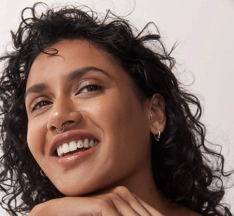 POETRY Skincare. Woman with brown curly hair, brown eyes, brown skin, nose ring smiling lookingnup to her left holding a POETRY Skincare Nourishing Cleansing Oil 100ml purple bottle with white pump in  her hands