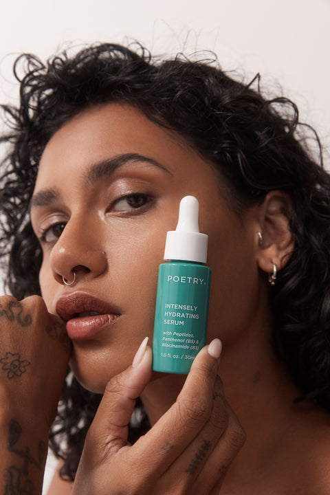 Woman with brown curly hair, brown eyes, nose ring, resting her hand with tattoos on it against the side of her face, holding a green POETRY Skincare bottle called Intensely Hydrating Serum against her face. 30ml bottle with white dropper with Vitamin B3, B5, hyaluronic acid and peptides.