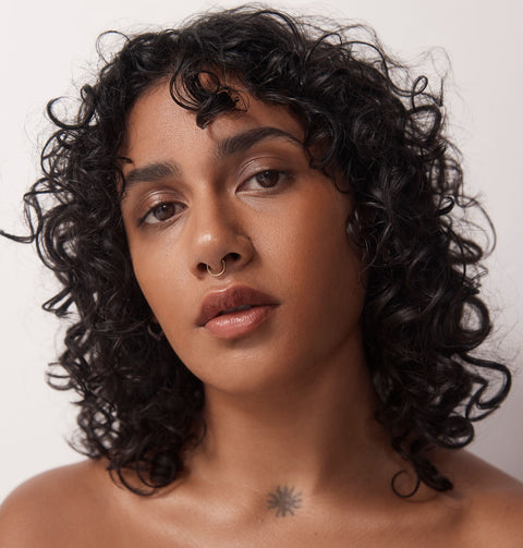 Poetry Skincare. Woman with brown curly hair worn down, brown eyes, brown skin, nose ring, mouth closed starring at the camera. No makeup