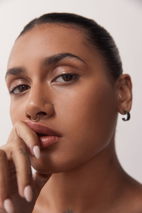 Woman with dark brown hair pulled back, staring at the camera with bent finger pressed up against her top lip pulling it up. She has a gold nose ring, no makeup, brown eyes, brown skin.