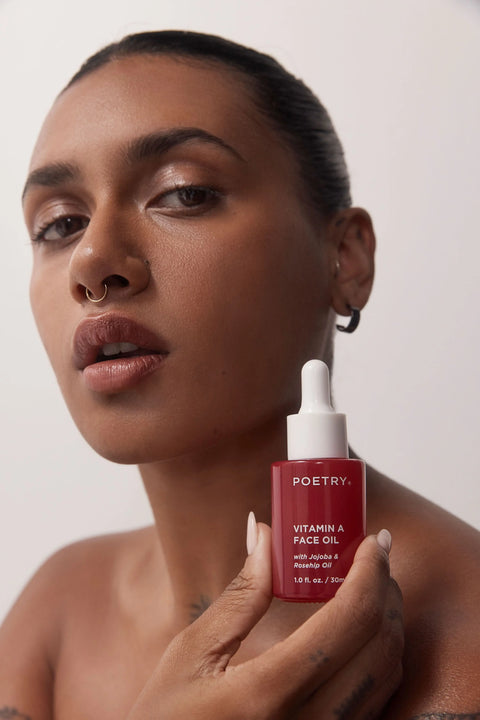Woman with brown hair pulled back, brown eyes, brown skin, medium sized looped earrings starring at the camera with mouth slightly open holding up a POETRY Skincare Vitamin A Face Oil 30ml glass bottle with white writing and white dropper with organic Rosehip and organic Jojoba.